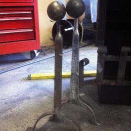 A close up of swan neck dog irons with ball tops and horse shoe feet. The one on the left is hand forged, the one on the right flame cut meaning its much